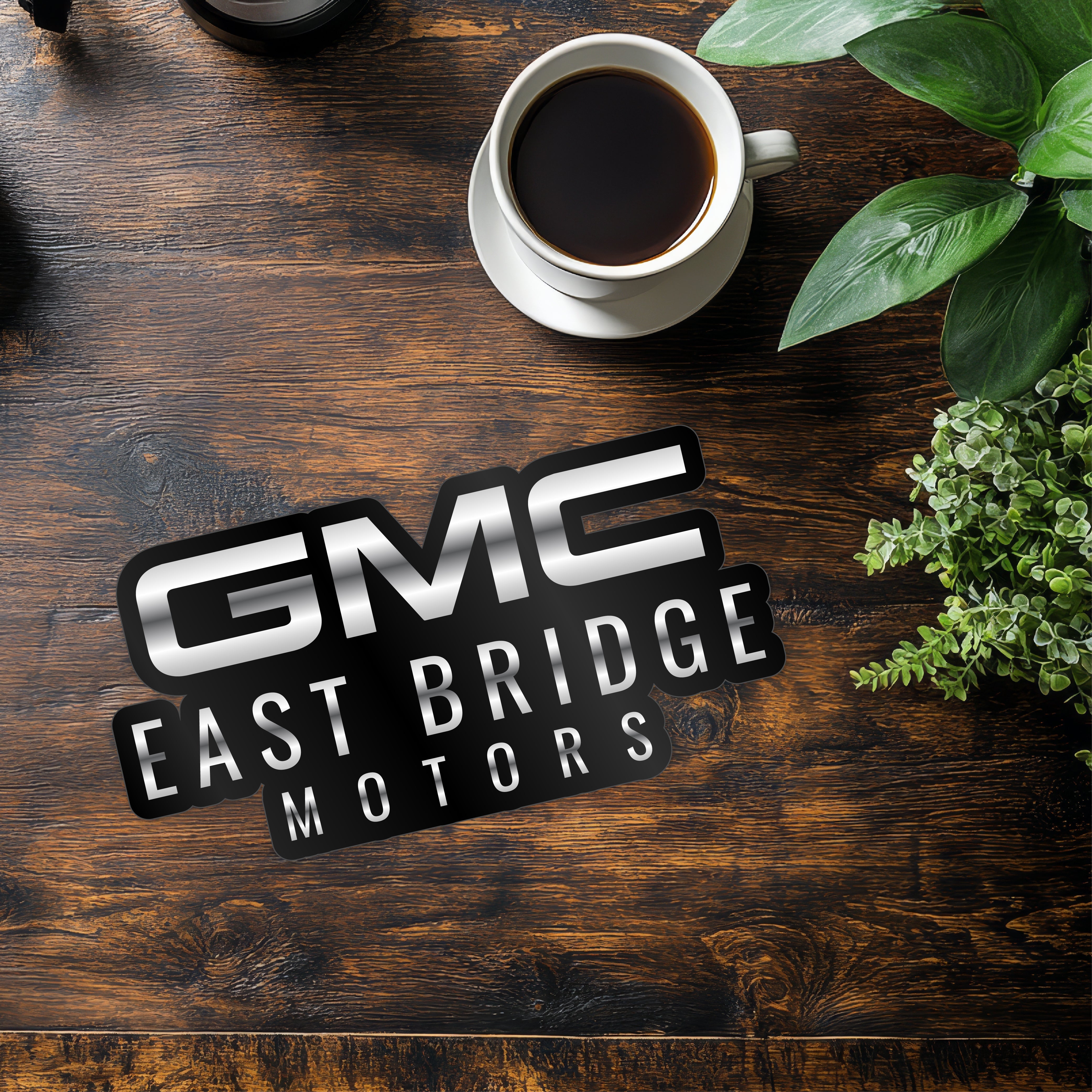 GMC East Bridge Motors logo displayed on a wooden table with coffee and plants, representing car dealership branding.